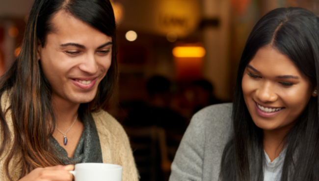 Two women sit in a cafe drinking coffee and looking at a laptop